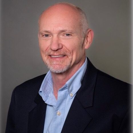 Professional headshot of a smiling bald man in a suit jacket and light blue shirt.